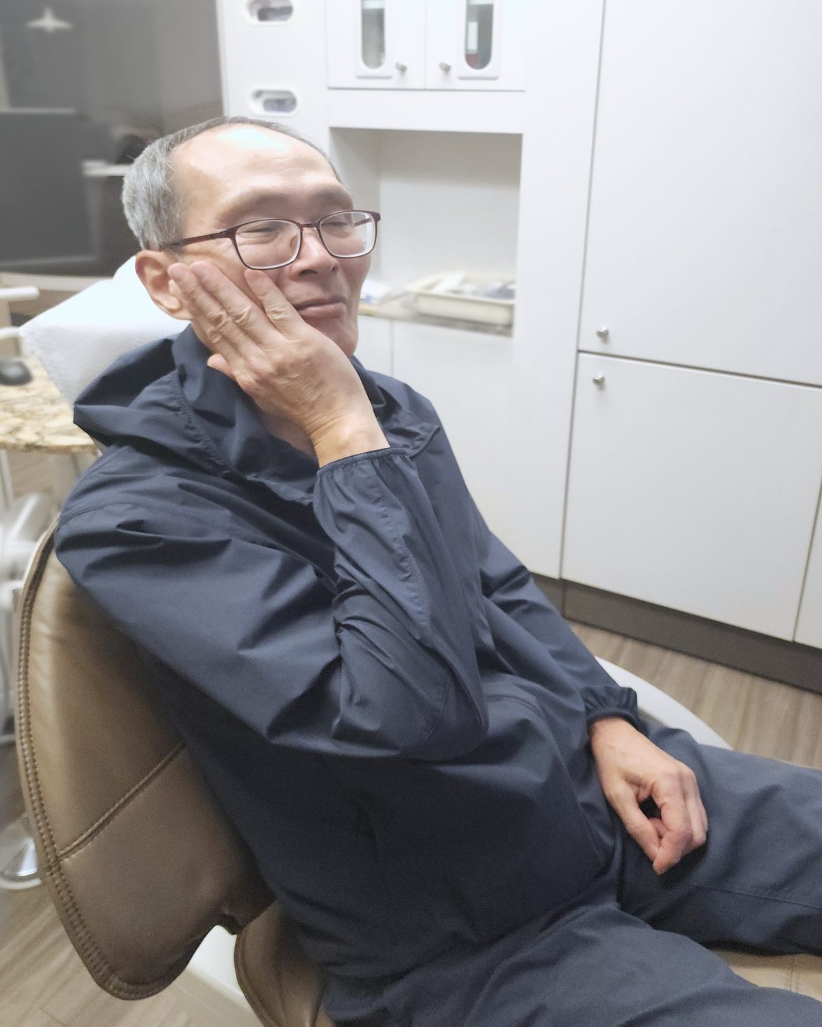 A man in a dental chair holding his cheek, as if he has a toothache.